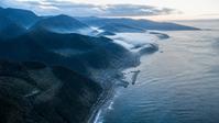 霧のかかる山並みと海沿いの町を上空から望む
